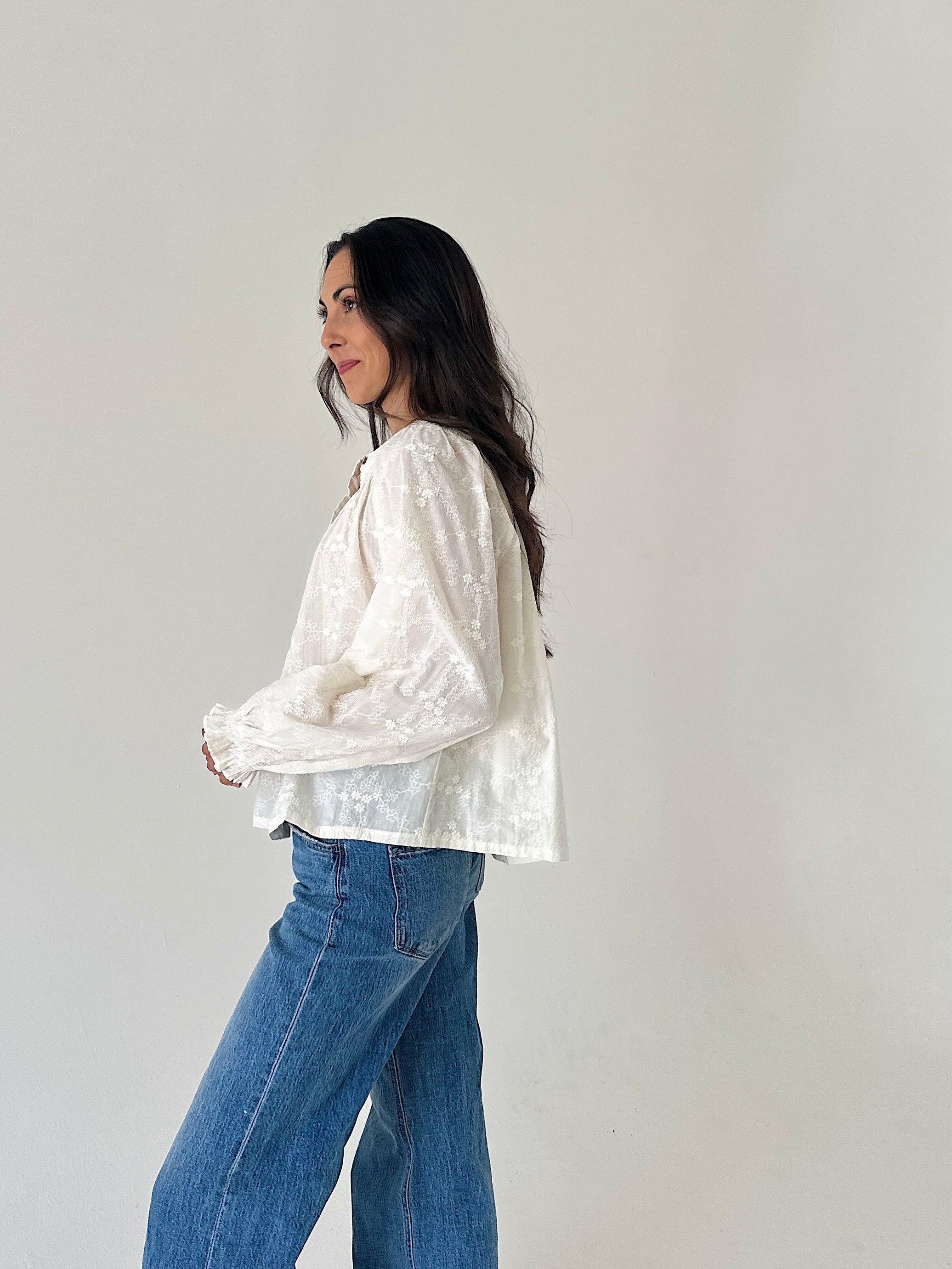 A woman wearing a white textured long-sleeve blouse and blue jeans, standing in profile against a plain light gray wall.
