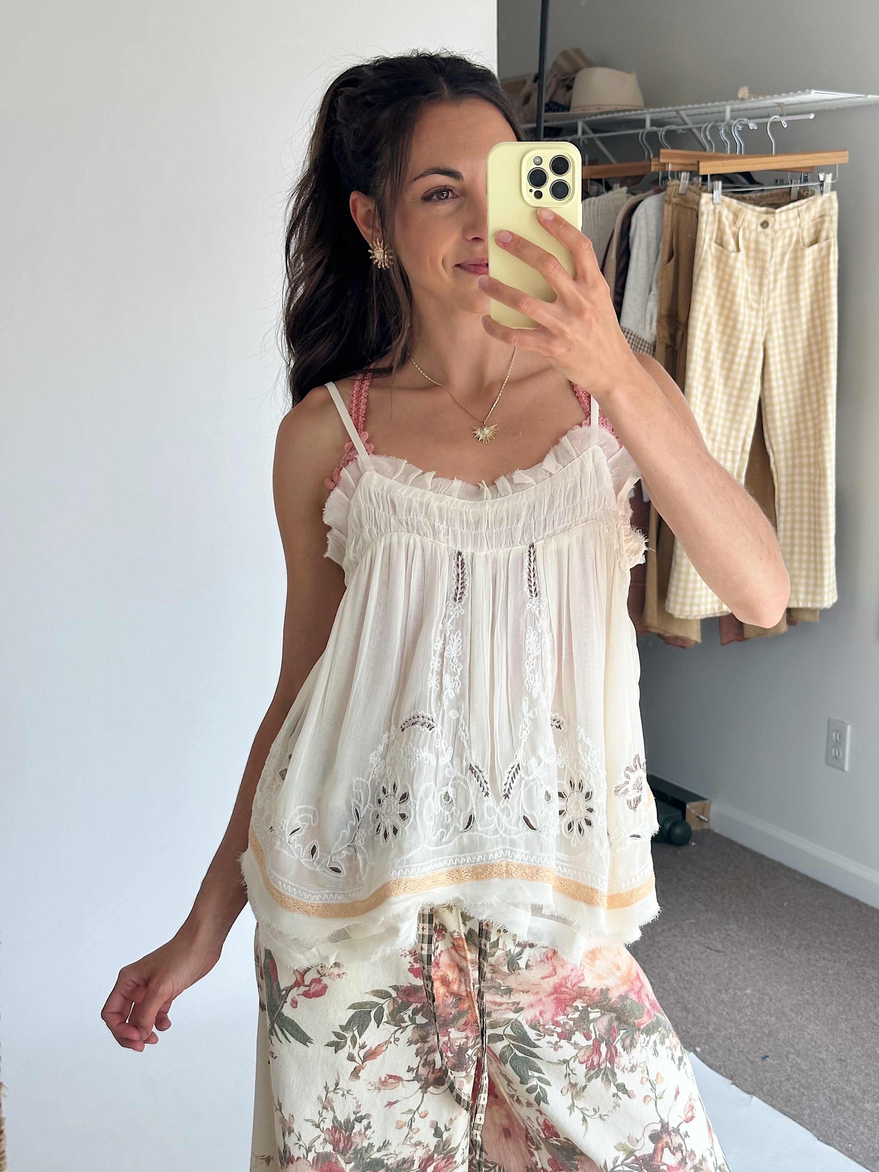 A woman takes a mirror selfie wearing a cream ruffled camisole top with floral embroidery and a floral-printed pants.