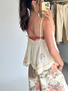 A woman takes a mirror selfie showing the back of a white lace camisole top with a pink braided strap detail and floral wide-leg pants.