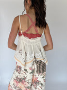 A woman shown from the back wearing a white lace-trim camisole top with a pink ruffled criss-cross strap design and floral print pants against a plain light gray background.
