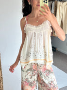 A woman takes a mirror selfie wearing a white ruffled camisole top with delicate embroidery and a floral print pants featuring pink, red, and green botanical patterns. She is holding a light yellow smartphone in front of her face.