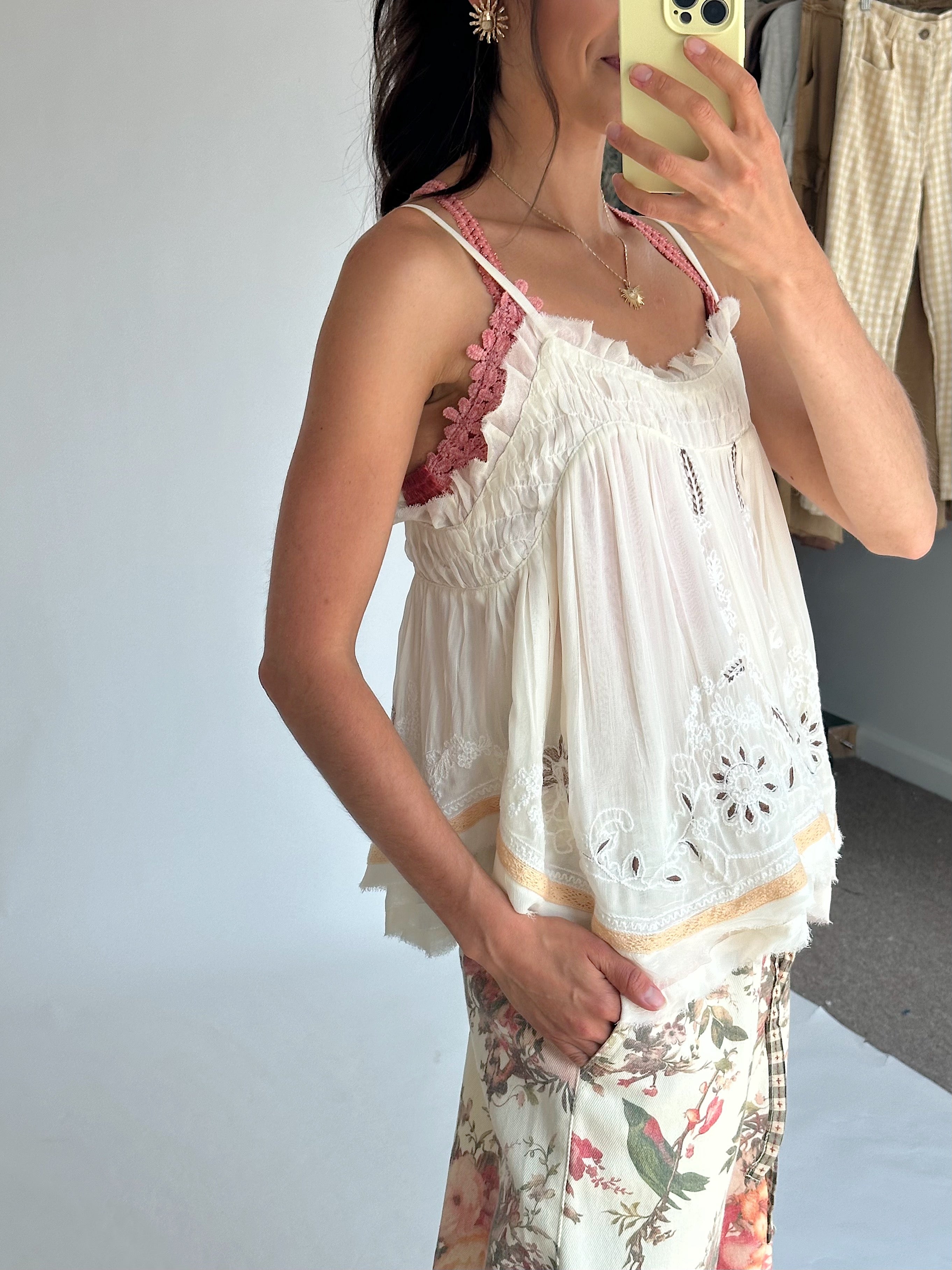 A woman takes a mirror selfie wearing a white eyelet lace camisole top over a pink bralette and floral print pants. She is holding a light yellow smartphone.