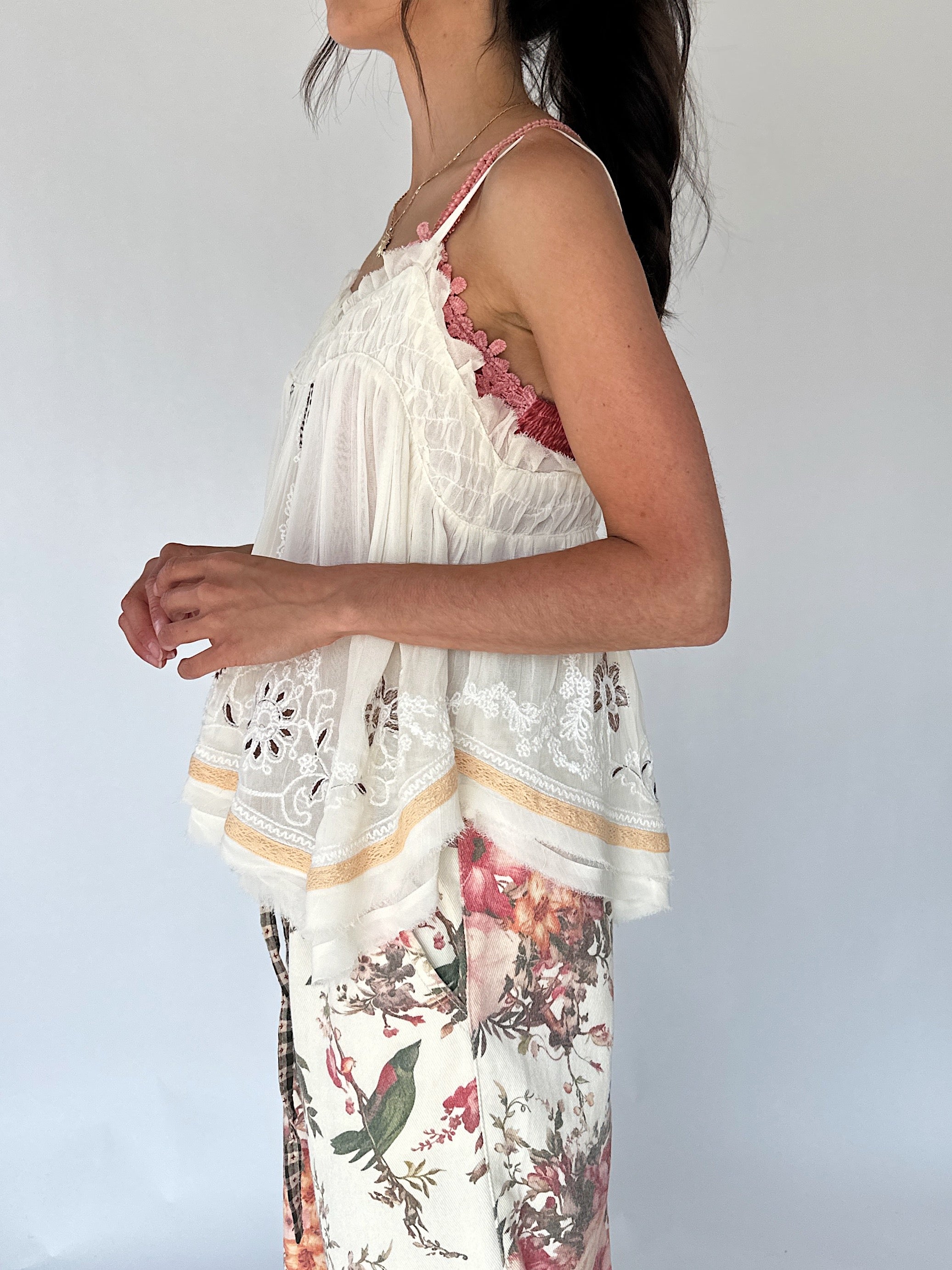 A woman shown from the side wearing a white camisole top with floral and lace details layered over a pink undergarment, paired with matching patterned bottoms, against a plain light gray background.
