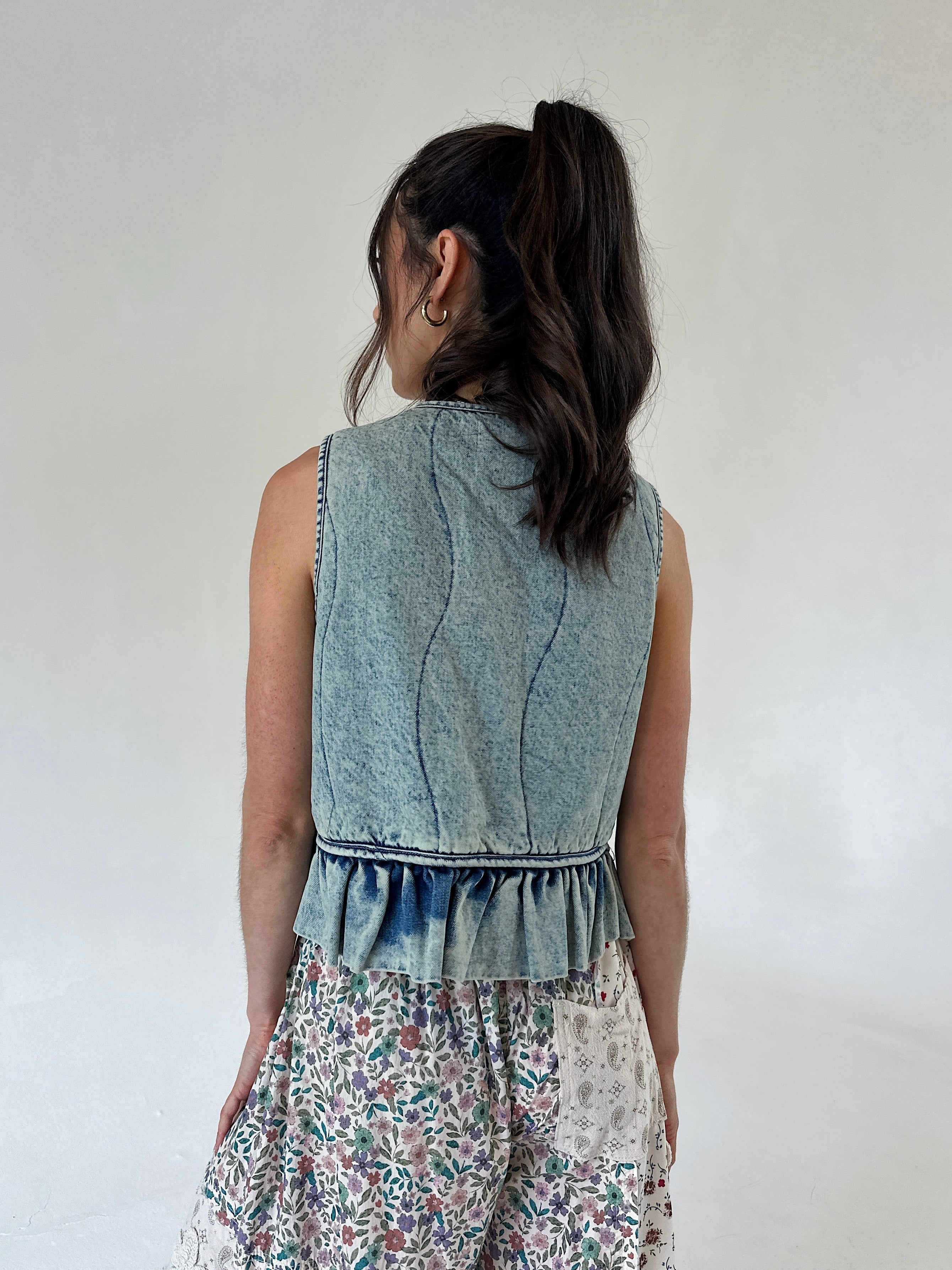 A woman shown from the back wearing a sleeveless light blue denim vest with a ruffled hem with floral pants, set against a plain off-white background.