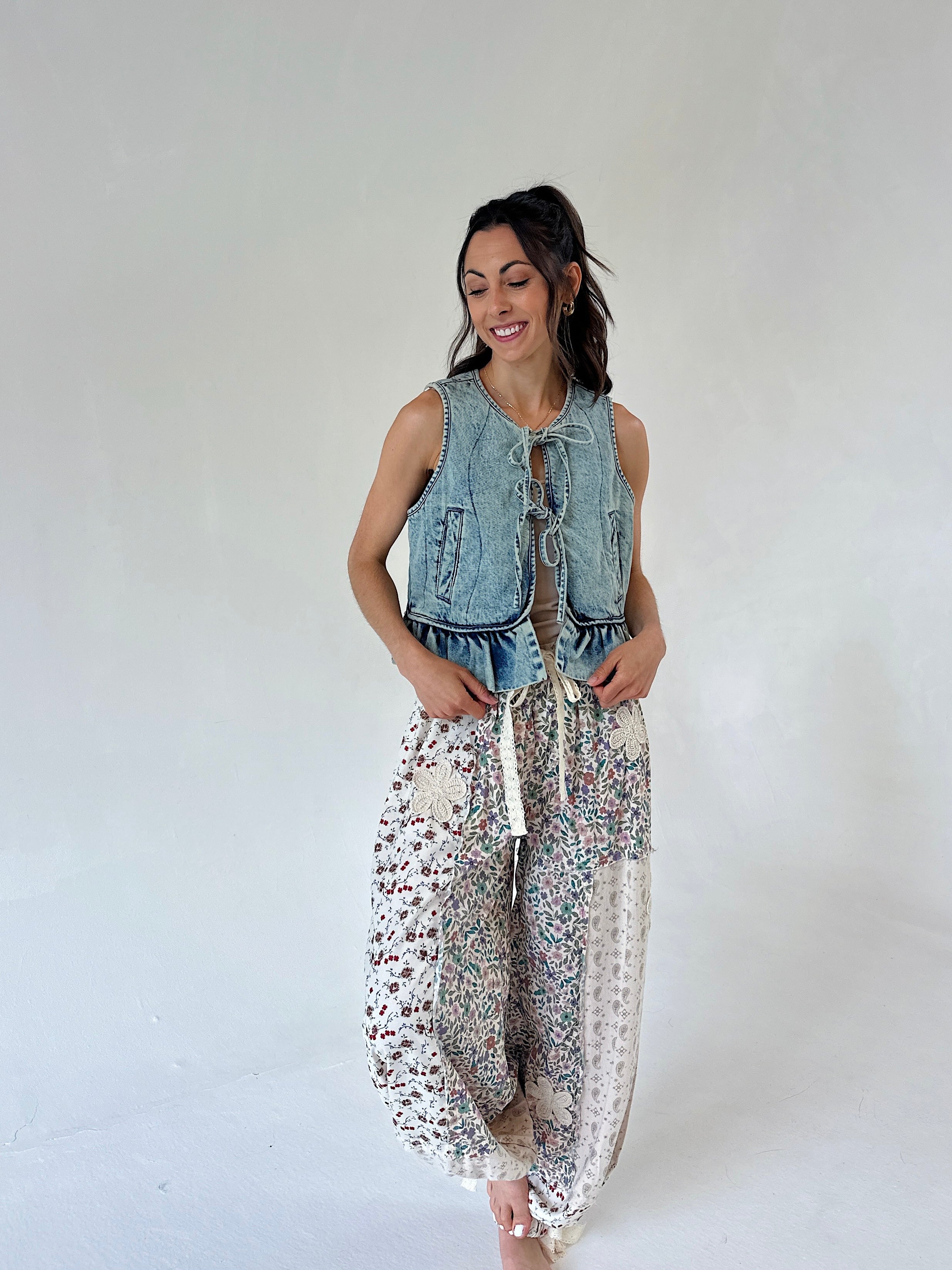 A woman modeling a sleeveless light-wash denim vest with front tie details and loose white harem-style pants covered in a small multicolor floral print, against a plain light gray studio background.