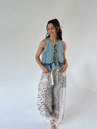 A woman modeling a sleeveless light-wash denim vest with front tie details and loose white harem-style pants covered in a small multicolor floral print, against a plain light gray studio background.