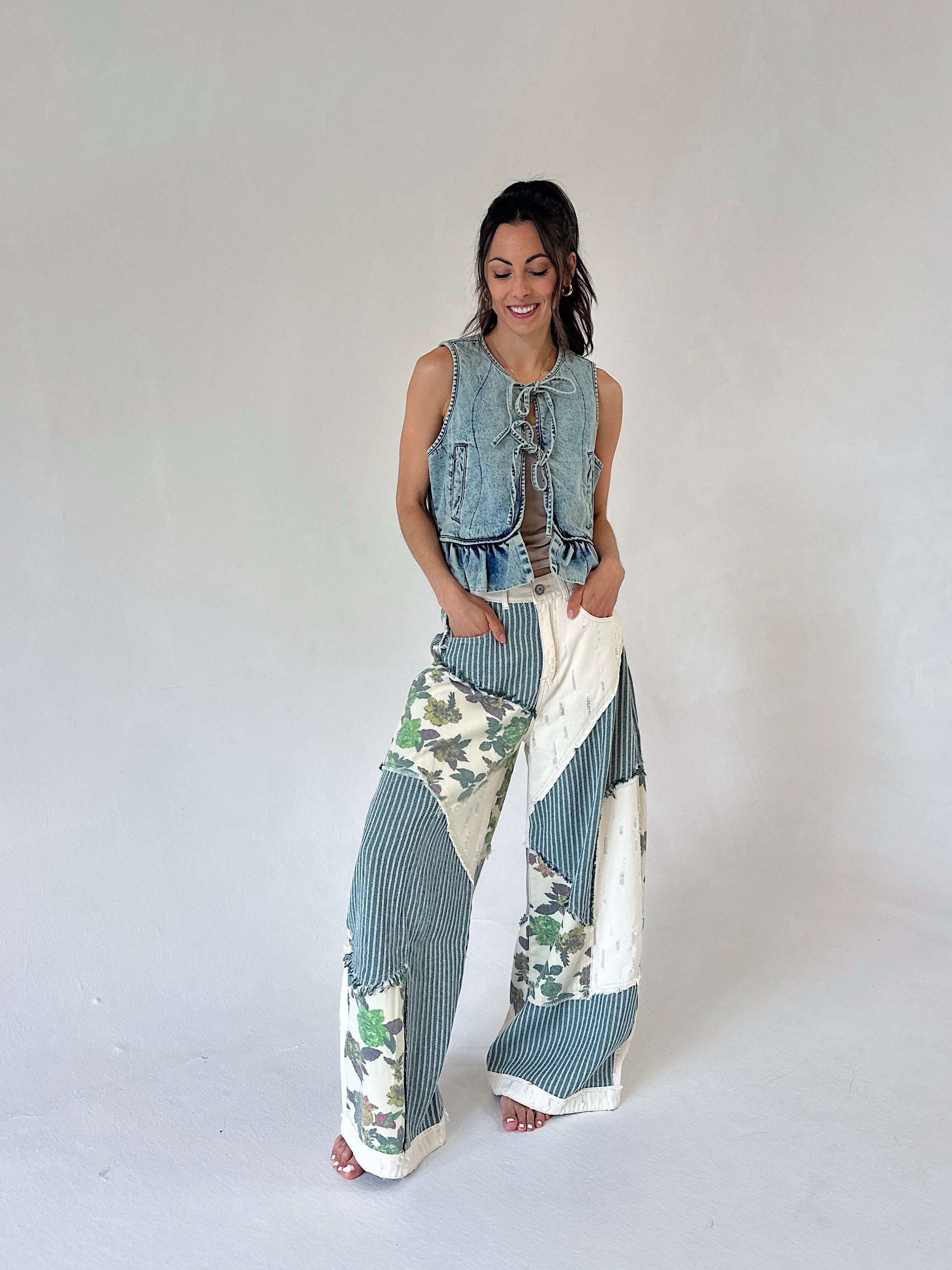 A woman modeling a sleeveless light blue denim vest paired with wide-leg patchwork pants featuring blue-gray and white stripes, floral prints, and solid white sections, against a plain light gray studio background.