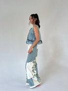 A woman stands in profile wearing a sleeveless denim vest with ruffled trim and wide-leg patchwork pants featuring blue stripes and white floral sections, against a plain light gray studio background.