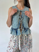 A woman wearing a sleeveless light-wash denim vest with tie-front detailing and ruffled hem, paired with floral print pants, against a plain light background.