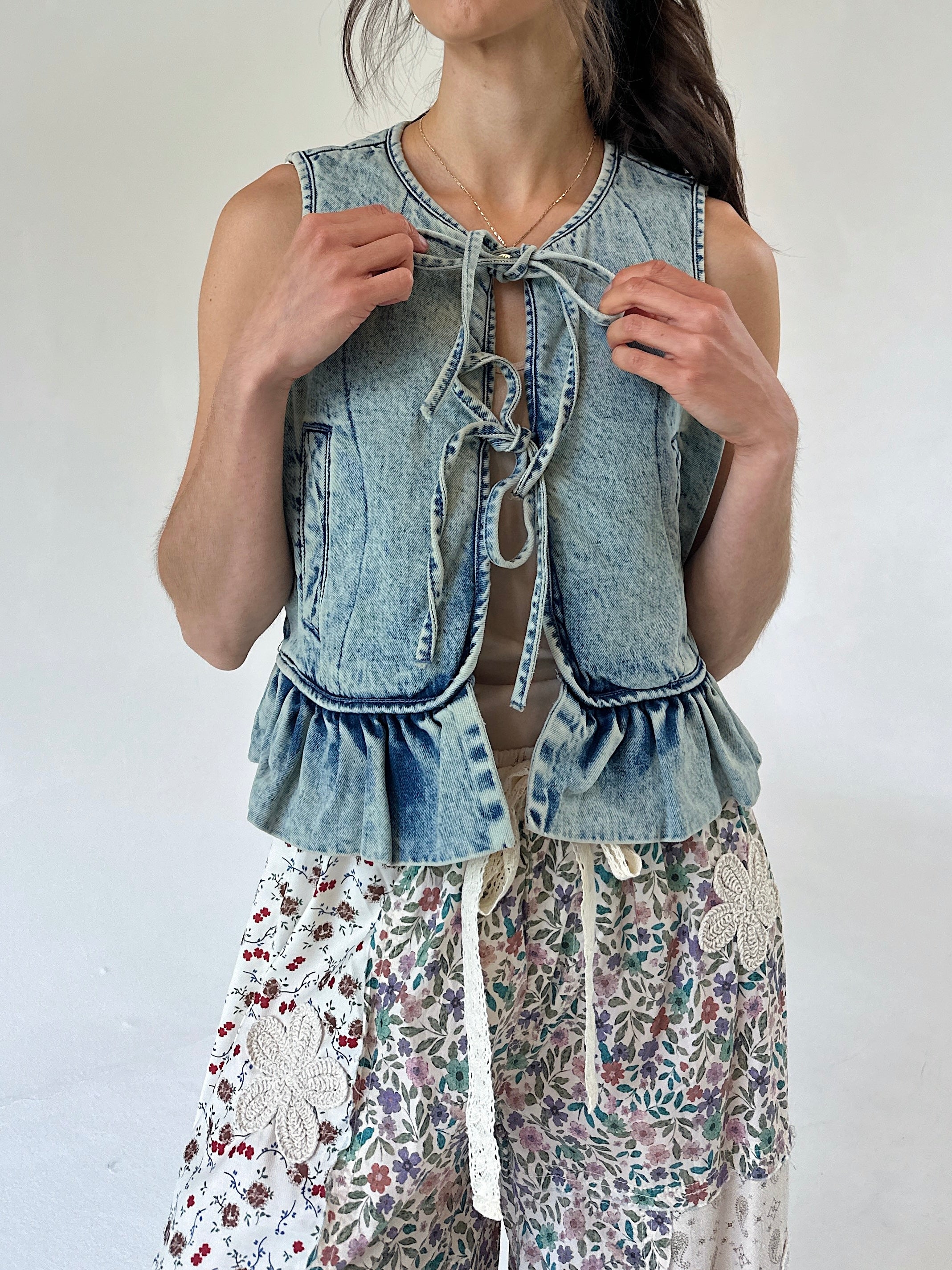 A woman wearing a sleeveless light-wash denim vest with tie-front detailing and ruffled hem, paired with floral print pants, against a plain light background.