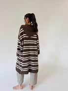 A woman stands barefoot against a plain off-white wall, viewed from the back, wearing a brown and cream horizontally striped hooded cardigan and brown-and-white checkered pajama pants.