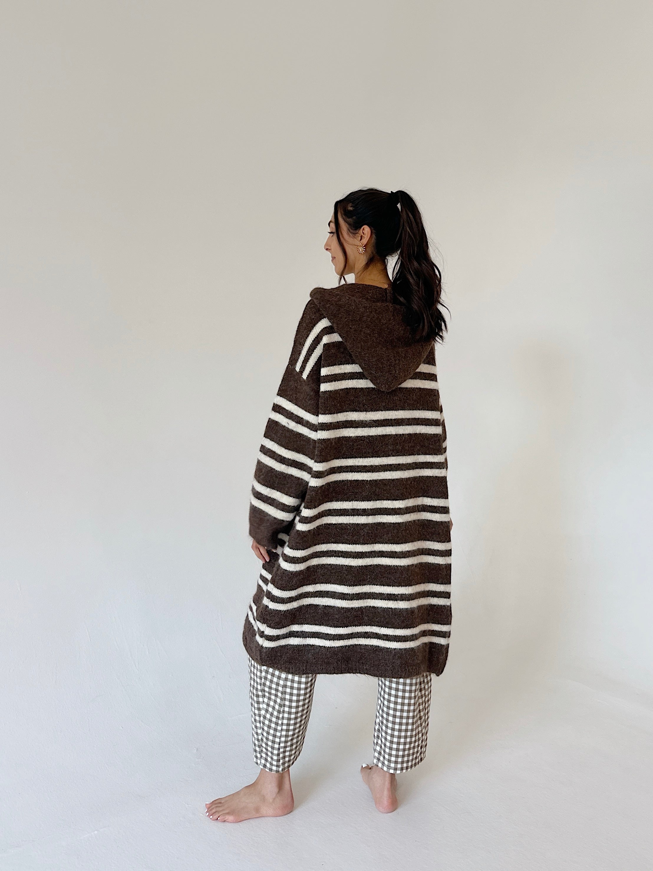 A woman stands barefoot in a studio wearing a brown and cream striped hooded cardigan or poncho-style top, paired with gingham pants, photographed against a plain off-white background.