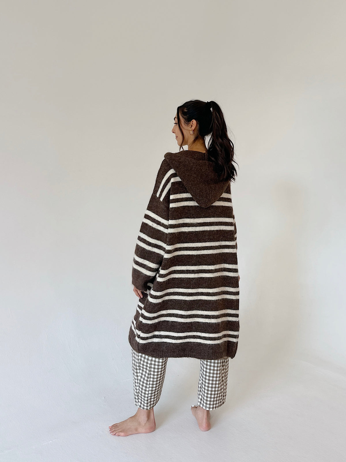 A woman stands barefoot in a studio wearing a brown and cream striped hooded cardigan or poncho-style top, paired with gingham pants, photographed against a plain off-white background.