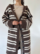 A person wearing a brown and cream striped fuzzy knit cardigan with a hood and front buttons, layered over a beige top, against a plain light background.