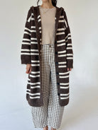 A person wearing a long brown and cream striped fuzzy cardigan over a beige top and brown-and-white gingham cropped pants, standing against a plain light background.