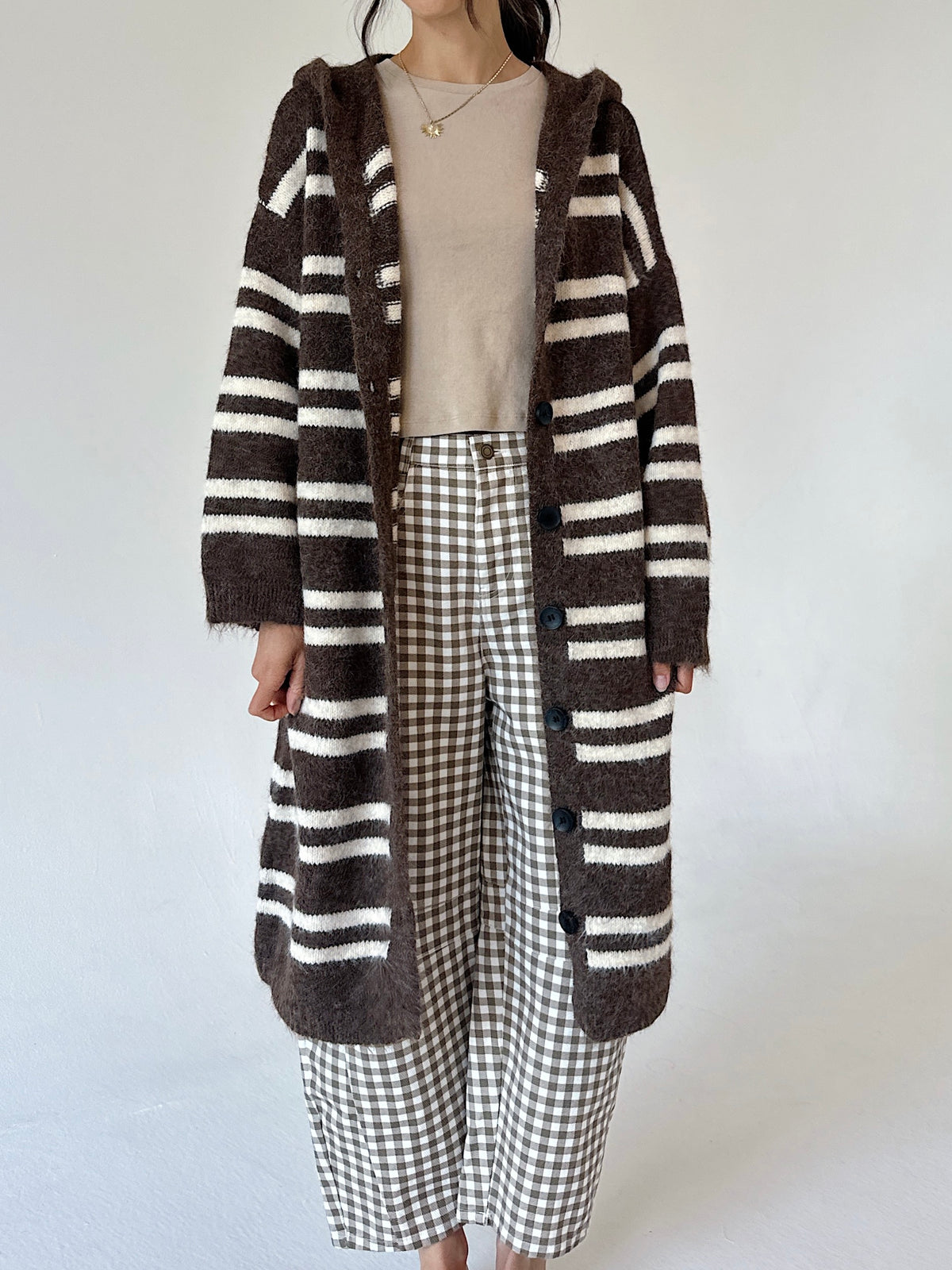A person wearing a long brown and cream striped fuzzy cardigan over a beige top and brown-and-white gingham cropped pants, standing against a plain light background.
