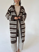 A woman wearing a long brown and cream striped knit cardigan over a beige top and brown-and-white gingham pants, standing against a plain light background.