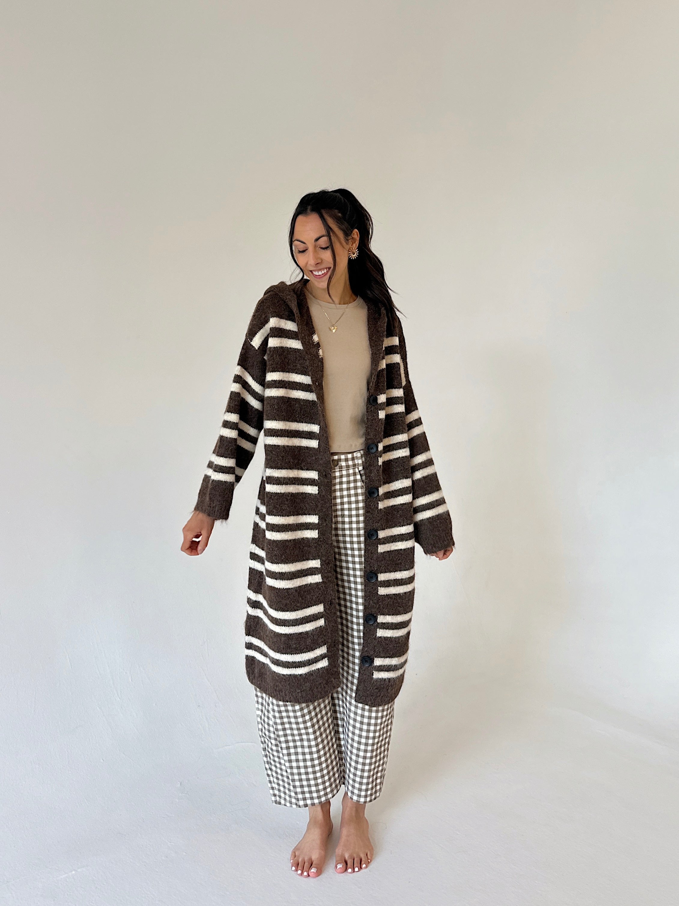 A woman stands barefoot against a plain light background wearing a long brown and cream striped fuzzy cardigan over a beige top and brown-and-white gingham cropped pants.