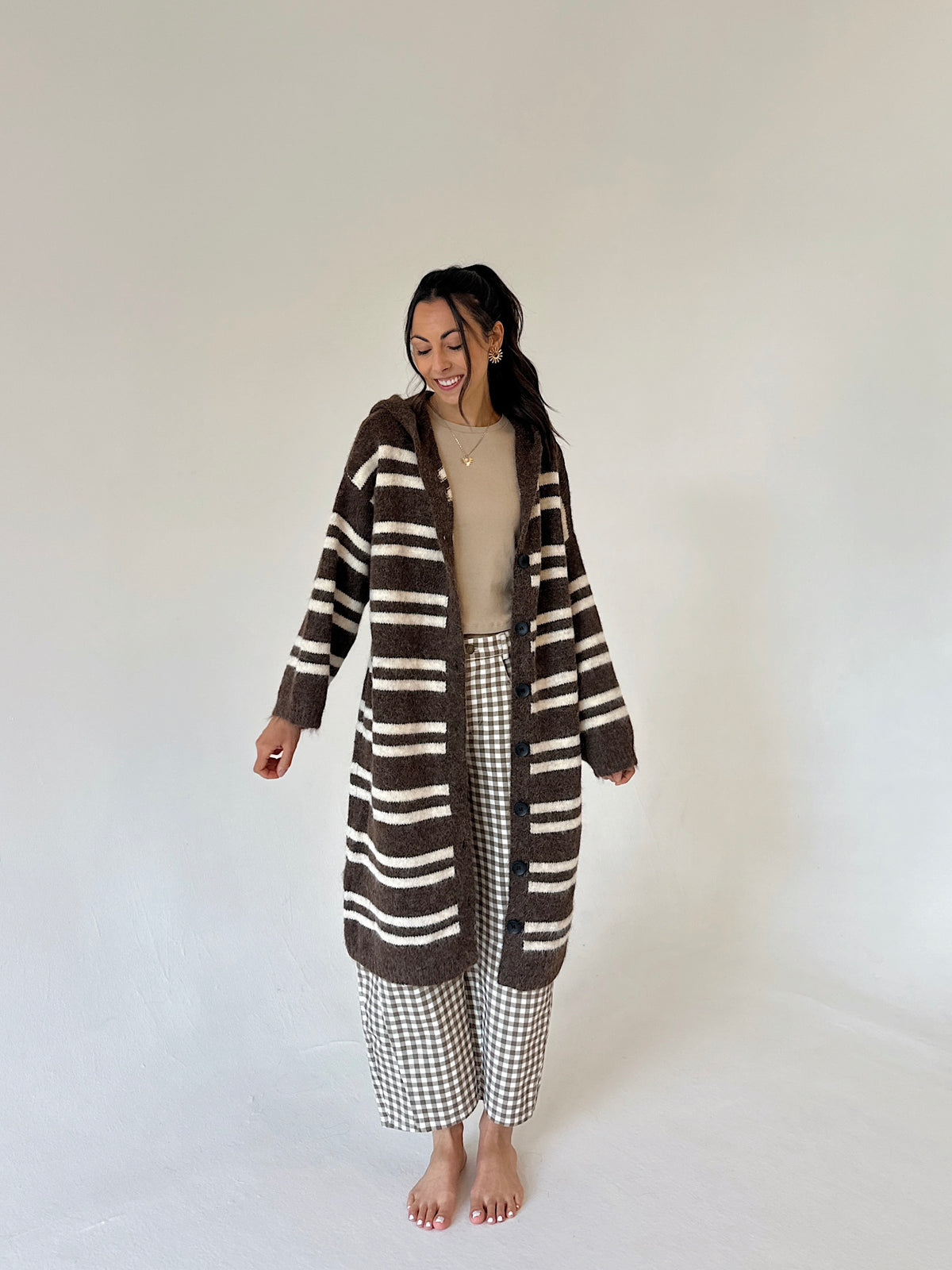 A woman stands barefoot against a plain light background wearing a long brown and cream striped fuzzy cardigan over a beige top and brown-and-white gingham cropped pants.