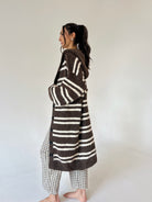 A woman stands barefoot in profile wearing a long brown and cream striped fuzzy cardigan over a brown-and-white gingham outfit, photographed against a plain light gray studio background.