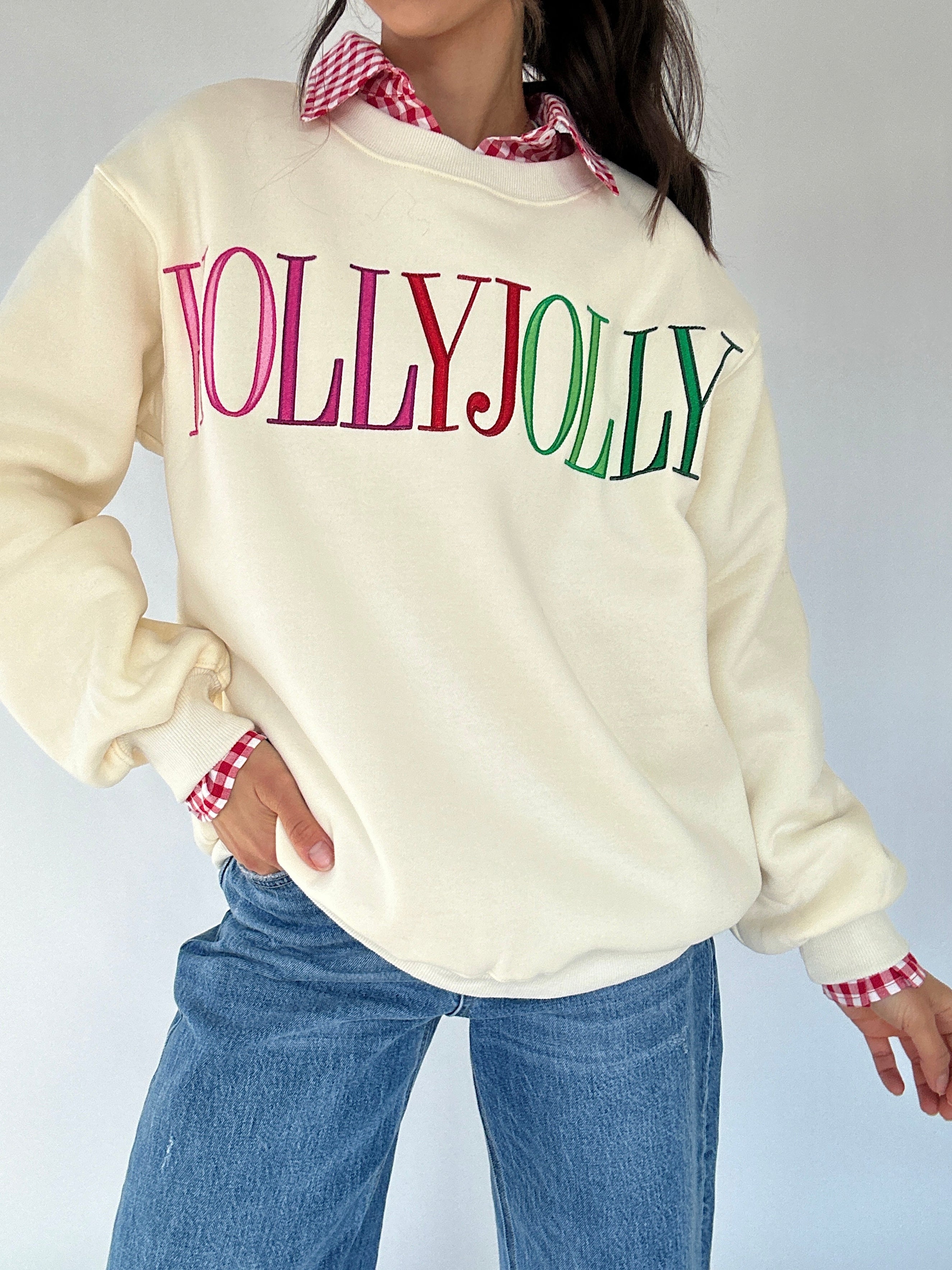 Person wearing a cream sweatshirt with 'HOLLY JOLLY' text, blue jeans, and checkered cuffs on a white background