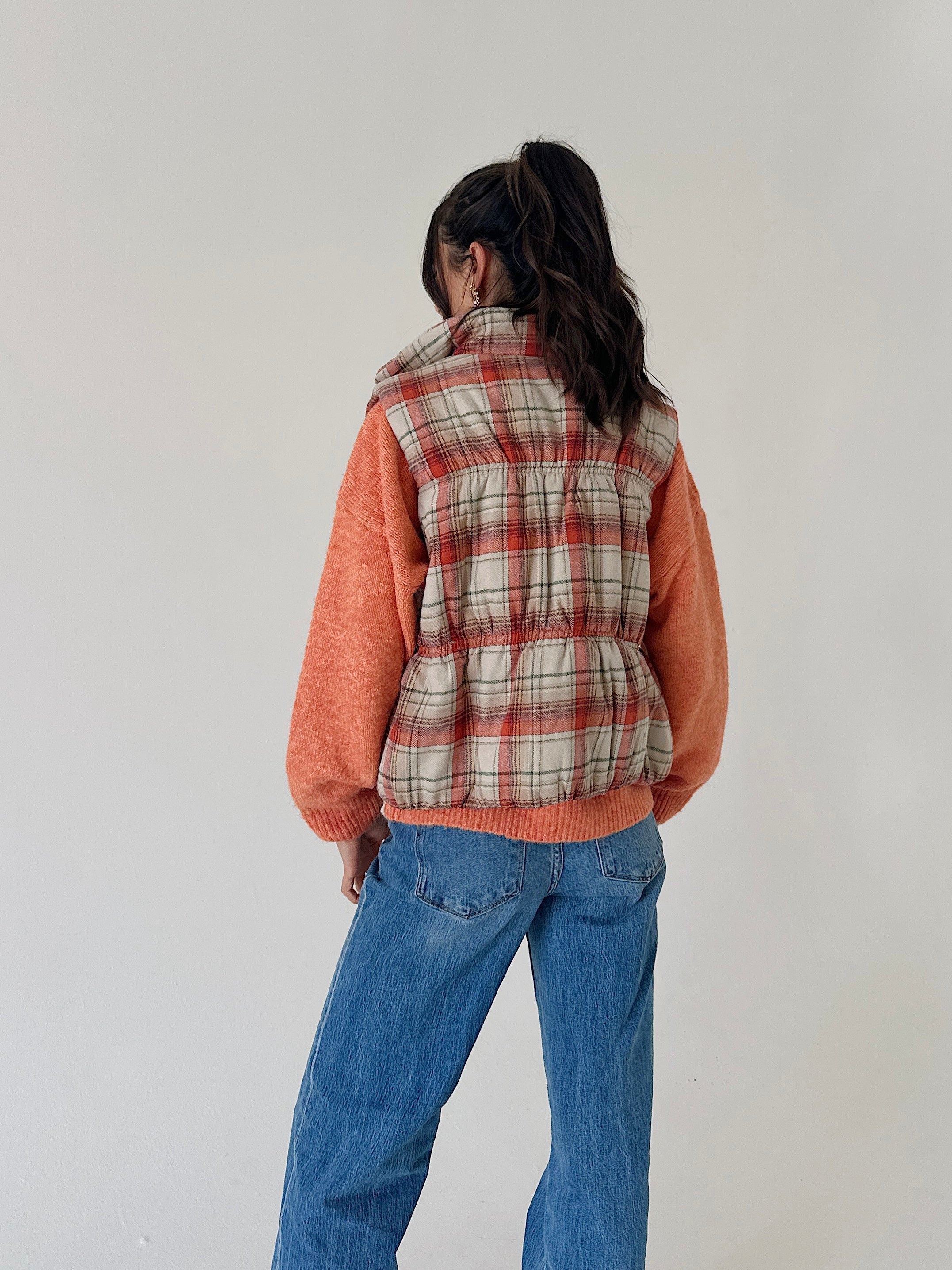 A person shown from the back wearing a sleeveless puffer vest with a beige, red, and white plaid pattern over a fuzzy orange sweater, paired with blue jeans, against a plain light gray wall.