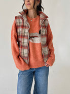 A woman wearing a textured orange knit sweater with a duck motif, layered under a red, beige, and white plaid zip-up vest, paired with blue jeans.