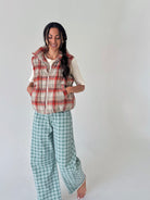 A woman modeling a plaid sleeveless puffer vest over a white ruffled shirt and green plaid wide-leg pants, standing against a plain light gray studio background.