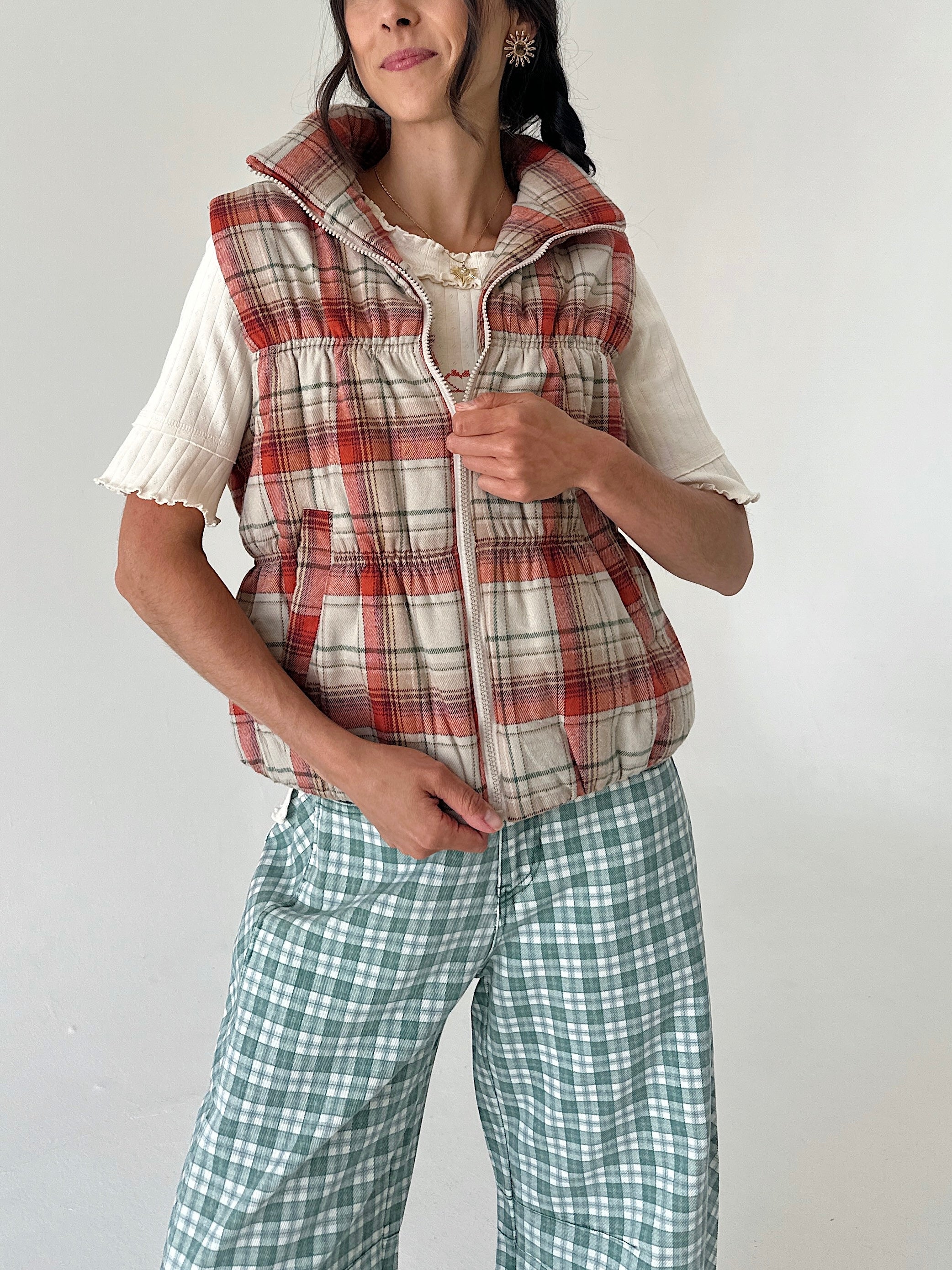 A woman wearing a sleeveless zip-front puffer vest in a red, beige, and white plaid pattern over a cream short-sleeve top, paired with green and white plaid pants, against a plain light gray background.