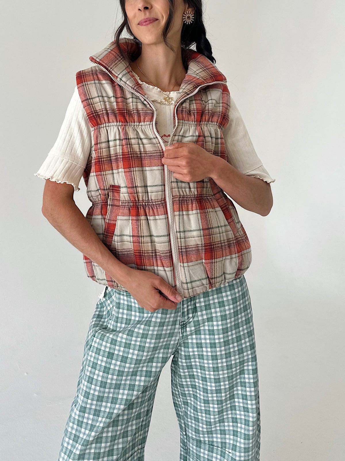 A woman wearing a sleeveless zip-front puffer vest in a red, beige, and white plaid pattern over a cream short-sleeve top, paired with green and white plaid pants, against a plain light gray background.