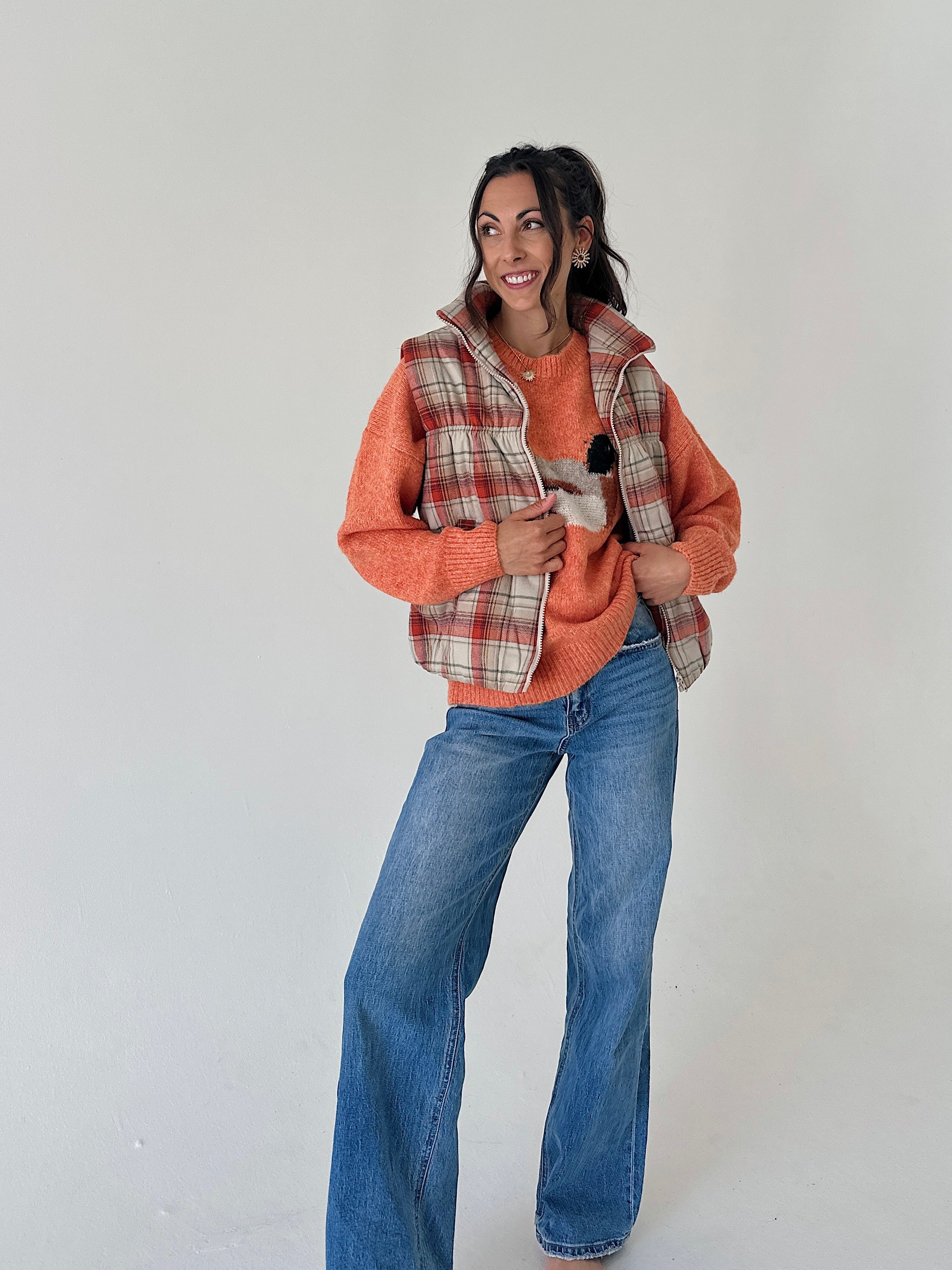 A woman modeling a rust-orange knit sweater vest layered over a plaid plaid hooded jacket, paired with blue jeans, against a plain light gray studio background.