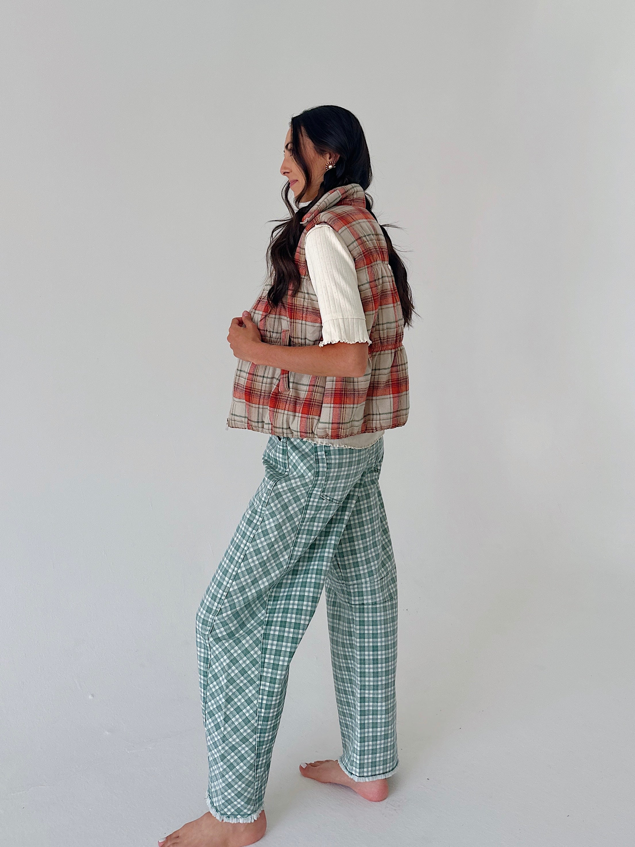 A woman stands barefoot in profile wearing a sleeveless red and beige plaid vest over a white short-sleeve top and green-and-white plaid pants, against a plain light gray studio background.