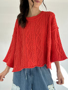 Person wearing a bright coral cable knit sweater and blue jeans on a white background