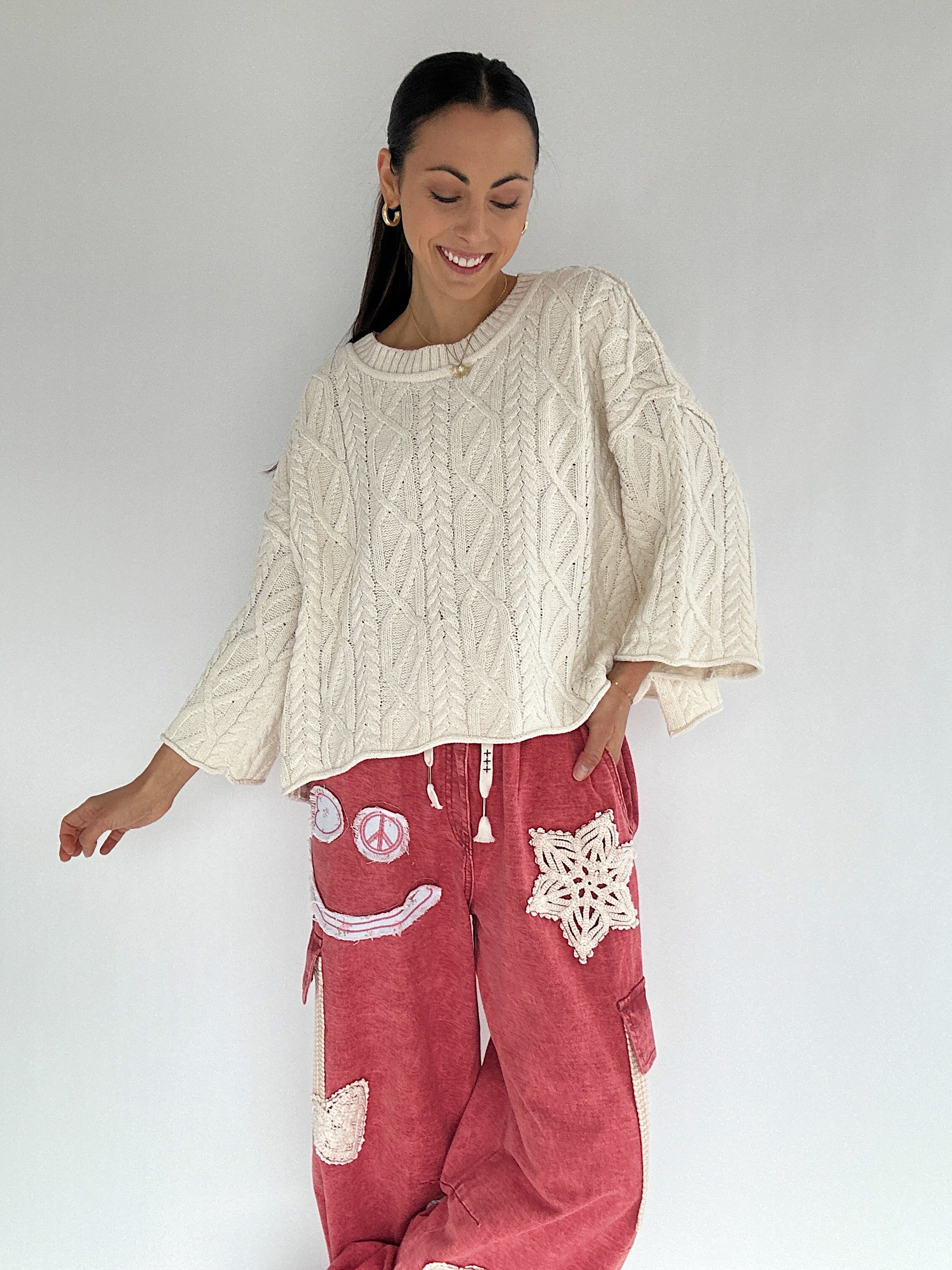 Woman wearing a cream cable knit sweater and red pants with patches on a white background