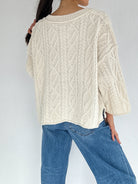Person wearing a cream cable knit sweater and blue jeans against a white background back view