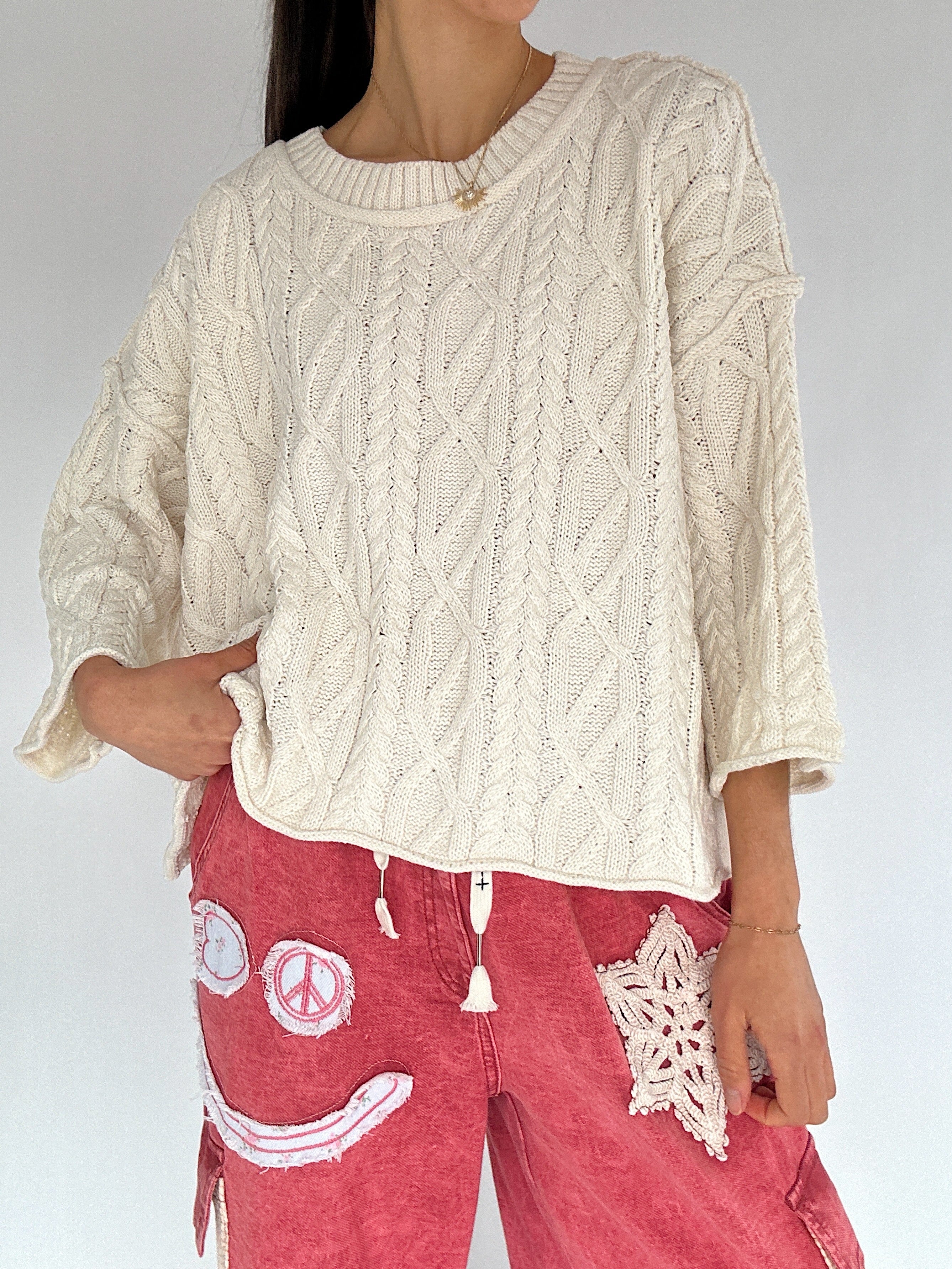 Person wearing a cream cable knit sweater and red pants with smiley face patch design.