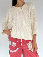 Person wearing a cream cable knit sweater and red pants with smiley face patch design.