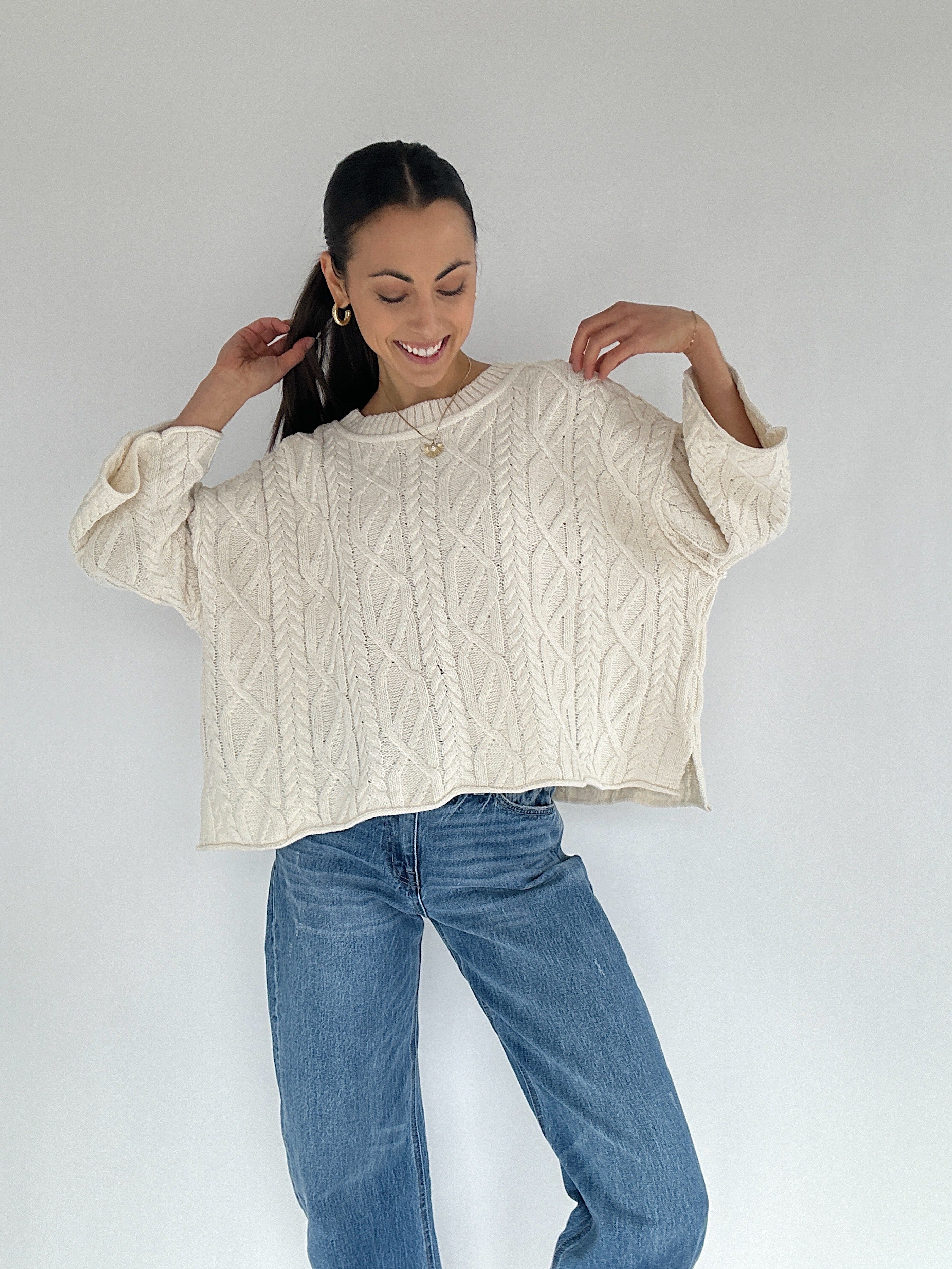 Person wearing a cream cable knit sweater and blue jeans against a white background