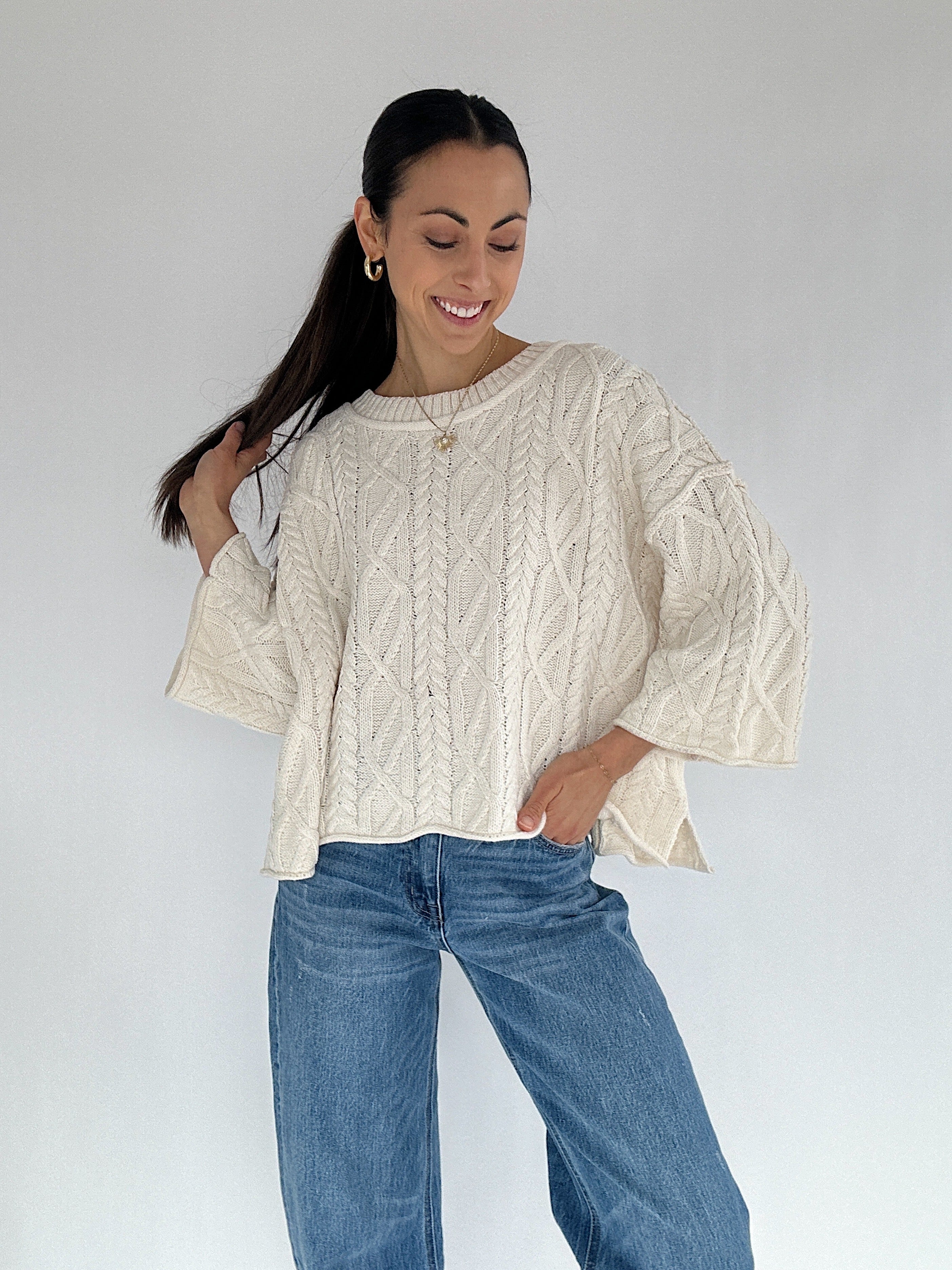 Woman wearing a cream cable knit sweater and blue jeans against a white background