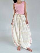 Person wearing a pink sleeveless top and white pants with lace and embroidery details on a plain background