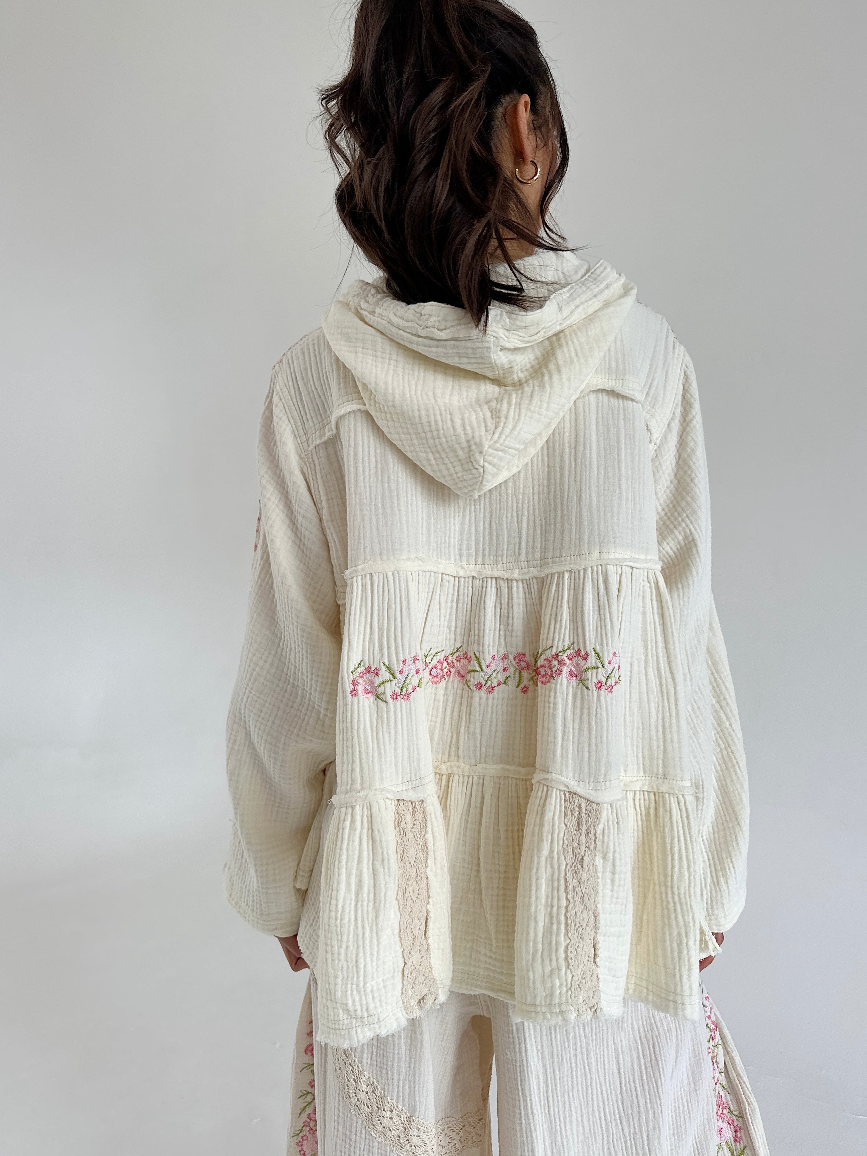 Woman wearing a white embroidered hooded top with a plain background showing the back