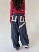 A person shown from the back wearing loose dark blue pants with large cream lace applique details and a pink long-sleeve top, standing against a plain light background.
