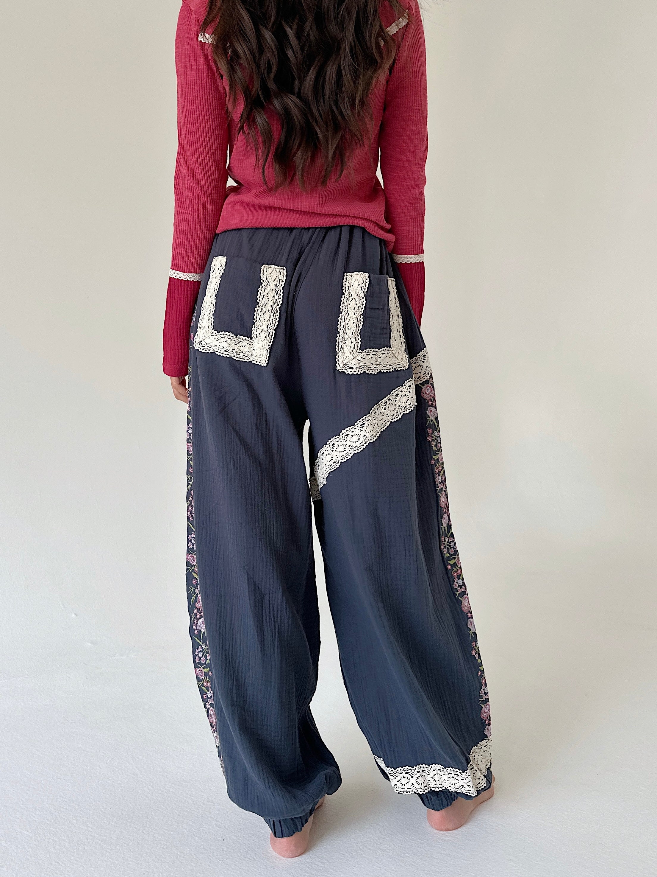 A person shown from the back wearing loose dark blue pants with large cream lace applique details and a pink long-sleeve top, standing against a plain light background.