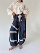 A model wearing a cream lace-trimmed long-sleeve blouse and navy blue gauze harem-style pants with white lace trim and a white drawstring tie, standing barefoot against a plain off-white studio background.