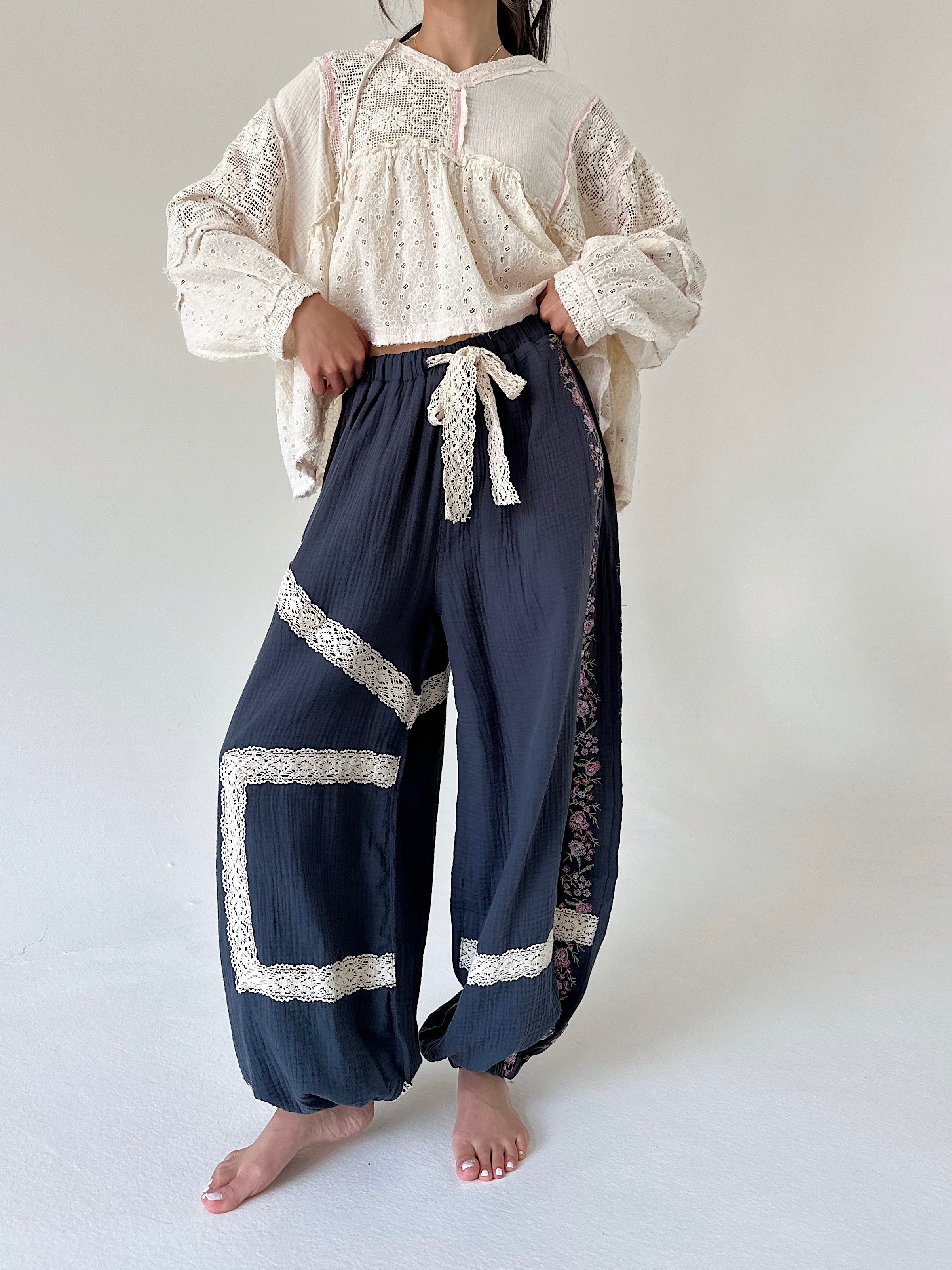 A model wearing a cream lace-trimmed long-sleeve blouse and navy blue gauze harem-style pants with white lace trim and a white drawstring tie, standing barefoot against a plain off-white studio background.