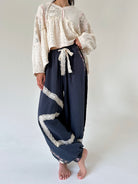 A model wearing a cream-colored lace long-sleeve crop top and dark navy blue crinkled wide-leg pants with white lace trim and a matching tie, standing barefoot against a plain light background.
