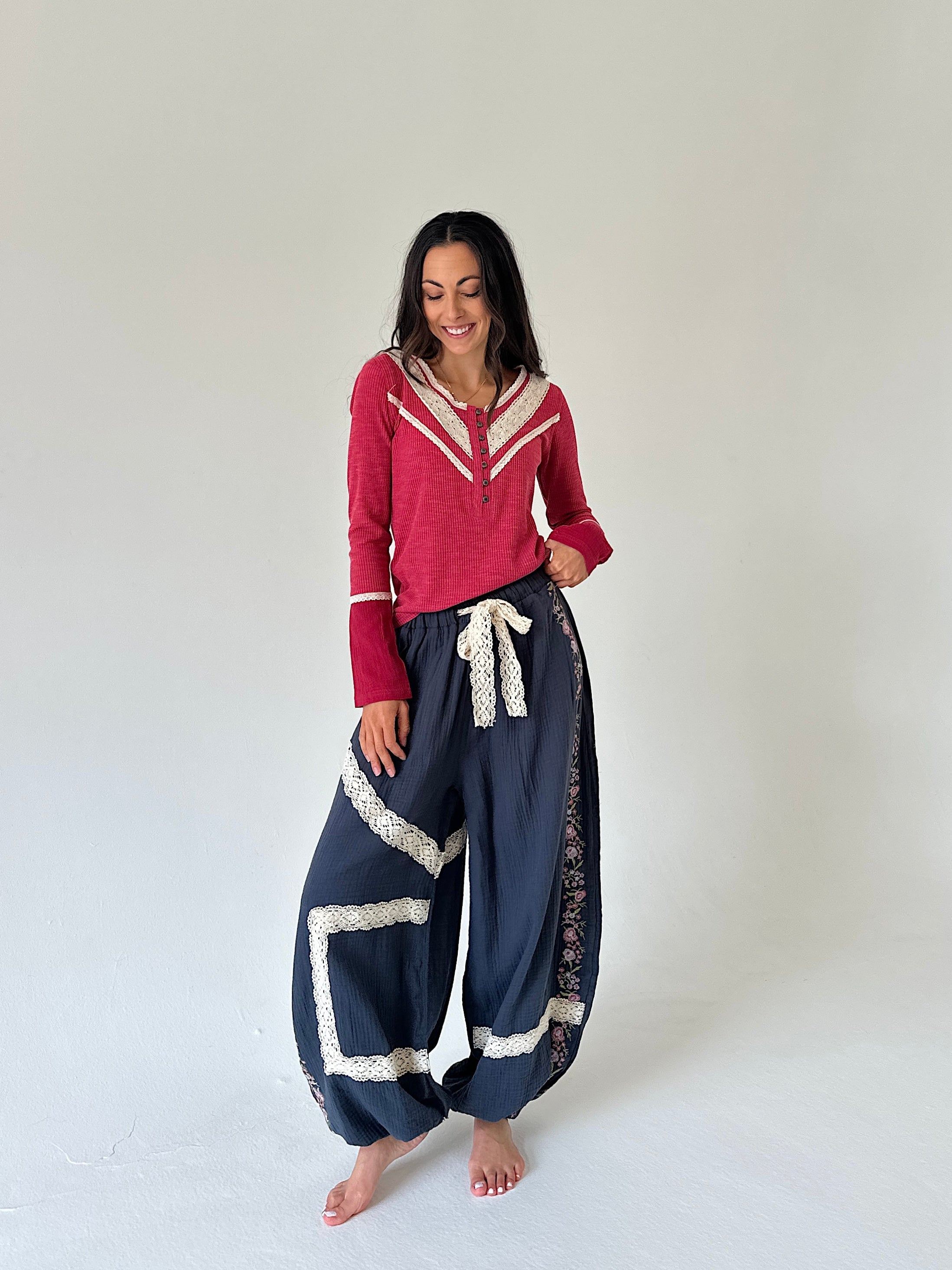 A woman modeling a red long-sleeve top with cream trim and navy blue wide-leg harem pants with cream lace trim, standing against a plain light gray studio background.