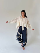 A woman modeling a bohemian-style outfit consisting of a cream lace long-sleeve blouse and navy blue harem pants with cream lace trim, standing against a neutral light gray studio background.