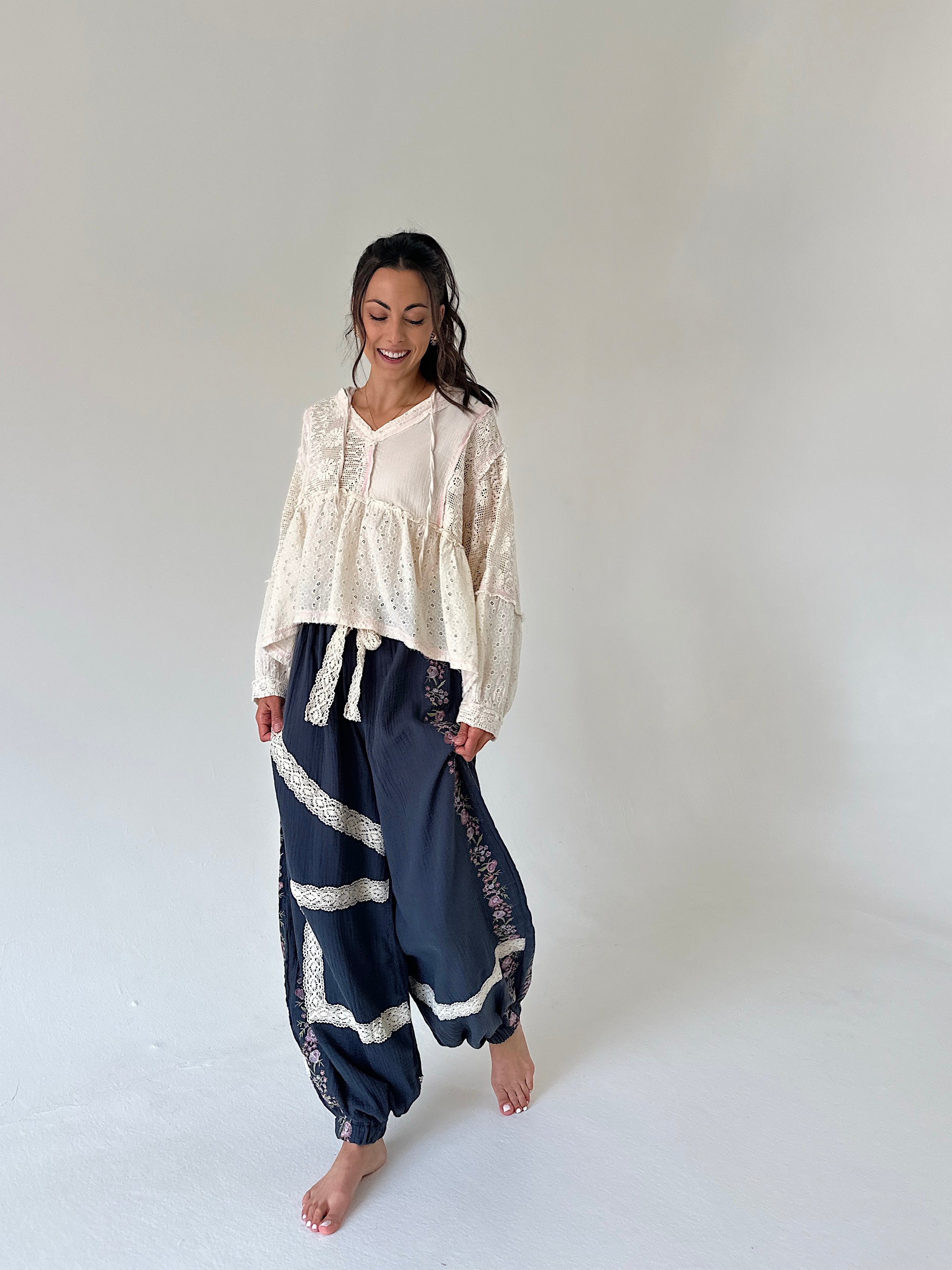 A woman modeling a bohemian-style outfit consisting of a cream lace long-sleeve top and navy blue patterned harem pants with lace trim, standing against a plain light gray studio background.