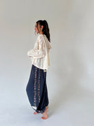 A woman stands barefoot in profile wearing a cream lace long-sleeve top and dark navy wide-leg pants with floral embroidered side panels, against a plain light gray studio background.